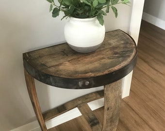 May include: A half-round wooden table with a metal band around the edge. The table is made from reclaimed wood and has a rustic look. A white ceramic planter with a green plant sits on the table.