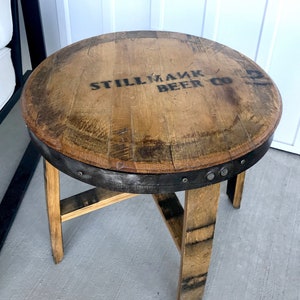 Może przedstawiać: Drewniany stolik z blatem z beczki i trzema nogami. Na blacie stolika widnieje napis "Stillmank Beer Co.".