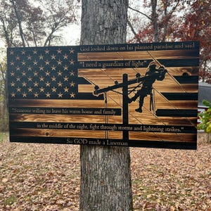 May include: A wooden American flag with a silhouette of a lineman working on a power line. The flag has a black background with white stars and red and white stripes. The text reads: "God looked down on his planned paradise and said "I need a guardian of light" "Someone willing to leave his warm home and family in the middle of the night, fight through storms and lightning strikes." So GOD made a Lineman"