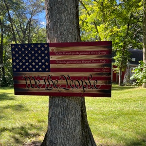 May include: A wooden sign with a distressed American flag design. The sign features the words "We the People" in a stylized font and a quote by Thomas Jefferson: "When the people fear the government, there is tyranny. When the government fears the people, there is liberty."