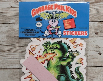 May include: A package of Garbage Pail Kids stickers featuring a green monster with a red tongue blowing fire. The package includes 5 stickers and 1 stick of bubble gum. The text on the package reads "Garbage Pail Kids", "All New! 2nd Series", and "Stickers".