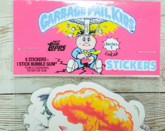 May include: A pack of Garbage Pail Kids stickers with a pink background. The pack includes 5 stickers and 1 stick of bubble gum. The stickers feature a cartoon character with a yellow explosion in their hair. The text on the pack reads "Garbage Pail Kids", "5 Stickers", "1 Stick Bubble Gum", "Not Topps", "Series 1", "Cheap", and "Stickers".