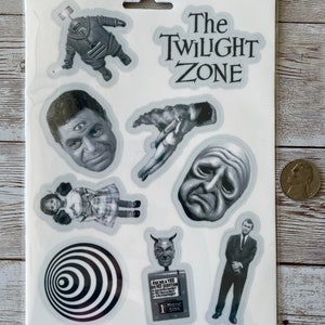 May include: A set of nine black and white stickers featuring characters and imagery from the Twilight Zone television series. The stickers include a robot, a man with three eyes, a hand, a crying face, a girl in a dress, a spiral, a devil, a man in a suit, and a sign that reads "Ask me a Yes or No Question".