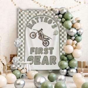 May include: A party decoration set featuring a banner with a motorcycle graphic and the text "MATTEO'S FIRST GEAR." The scene includes a balloon arch in silver, green, and cream colors, along with scattered balloons on the floor.