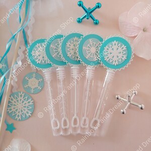 May include: Six blue and white snowflake-shaped bubble wands with clear plastic bottles filled with bubble solution. The wands are arranged in a row on a pink surface.