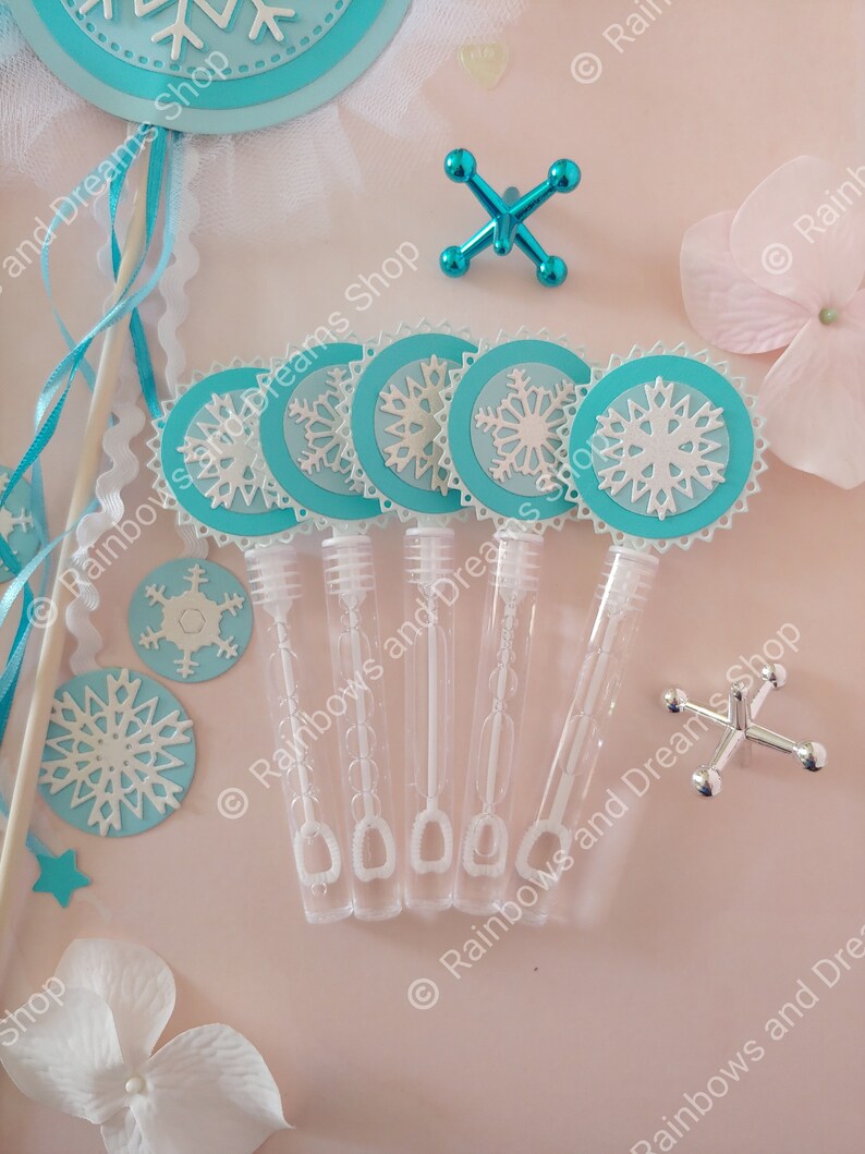 May include: Six blue and white snowflake-shaped bubble wands with clear plastic bottles filled with bubble solution. The wands are arranged in a row on a pink surface.