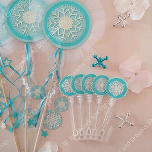 May include: A set of snowflake-themed party favors, including wands with blue and white snowflakes and clear plastic bubble wands with blue and white snowflake stickers. The favors are arranged on a pink surface with white flowers.