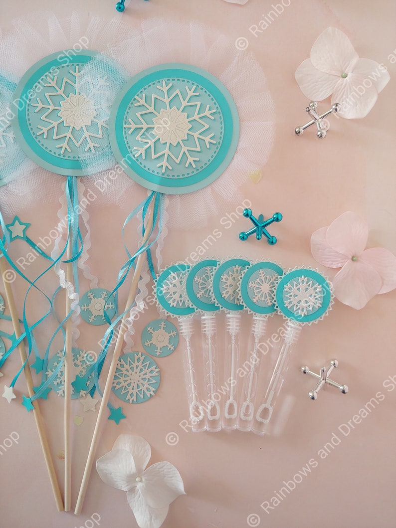 May include: A set of snowflake-themed party favors, including wands with blue and white snowflakes and clear plastic bubble wands with blue and white snowflake stickers. The favors are arranged on a pink surface with white flowers.