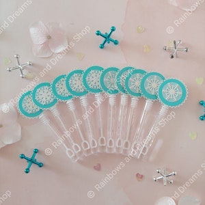 May include: Ten clear plastic bubble wands with turquoise snowflake-shaped toppers. The wands are arranged in a semi-circle on a pink surface with white flowers and silver and turquoise toy jacks.