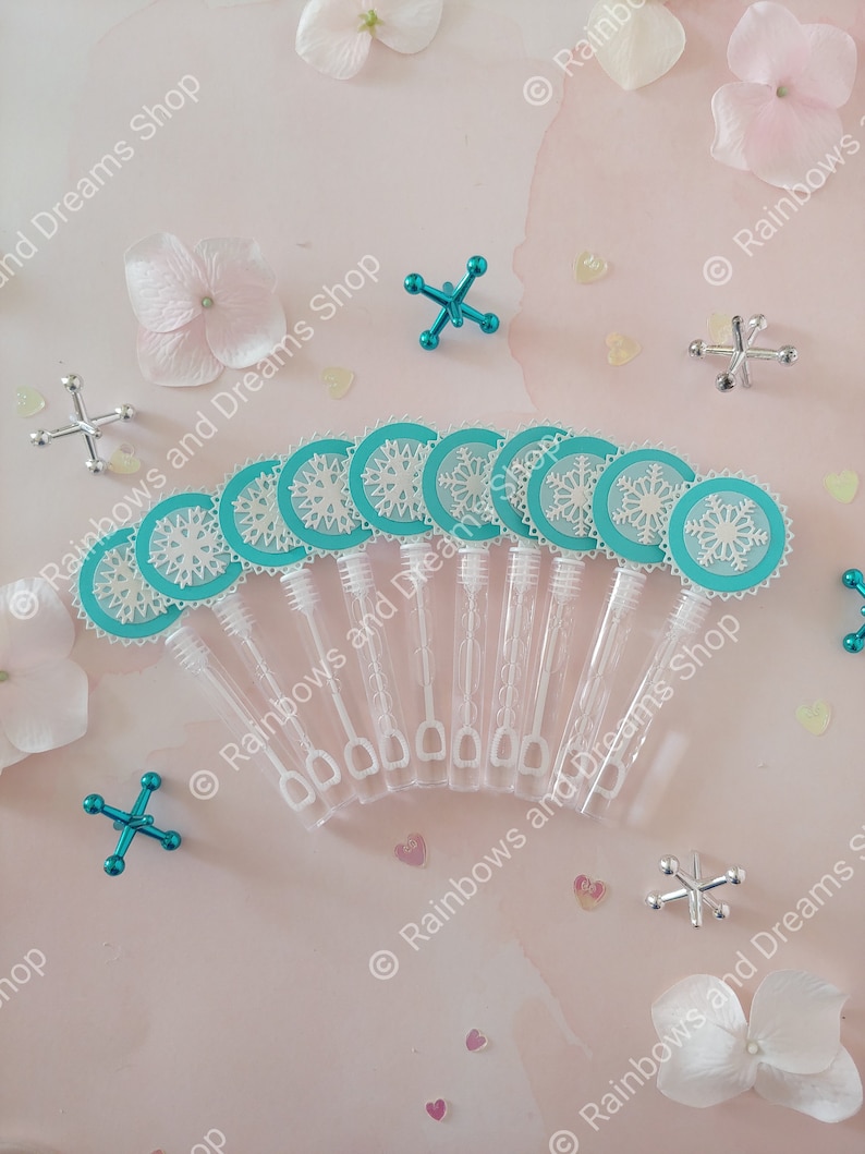 May include: Ten clear plastic bubble wands with turquoise snowflake-shaped toppers. The wands are arranged in a semi-circle on a pink surface with white flowers and silver and turquoise toy jacks.