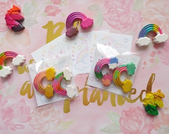 May include: A collection of colorful unicorn and rainbow-themed craft supplies. The image features rainbow-shaped wax crayons with white cloud accents, unicorn-shaped crayons, and small drawings of unicorns and rainbows. The background is a pink floral pattern.