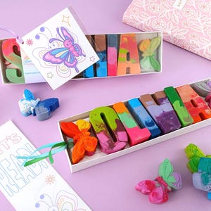 May include: A gift set featuring colorful crayons shaped into letters spelling a name, presented in a white box. The crayons are accompanied by butterfly-shaped wax figures and decorative tags with butterfly illustrations. The overall theme is a personalized gift.