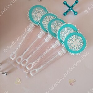 May include: Five clear plastic bubble wands with white handles and blue and white snowflake-shaped paper toppers. The wands are on a pink surface.