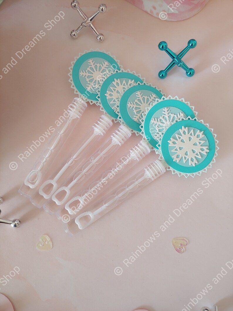 May include: Five clear plastic bubble wands with white handles and blue and white snowflake-shaped paper toppers. The wands are on a pink surface.