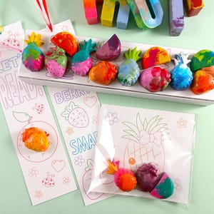May include: A collection of colorful fruit-shaped crayons, including pineapples, strawberries, and watermelon slices, arranged on a white tray. Two bookmarks with the text "LET READ" and "BERRY SMART" are also visible, along with a small bag of crayons.
