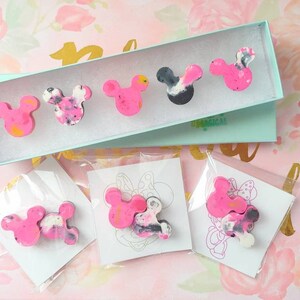 May include: A set of six pink and black resin Mickey Mouse head pins. The pins are packaged in a light blue box with the word "remarkable" printed on it. The pins are also packaged individually in clear plastic bags with white cards.