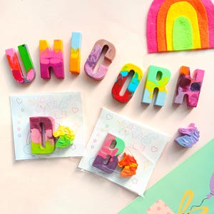 May include: A set of colorful crayon letters spelling out the word "UNICORN" with two additional crayons shaped like unicorns. The crayons are on a pink background with white paper packaging.