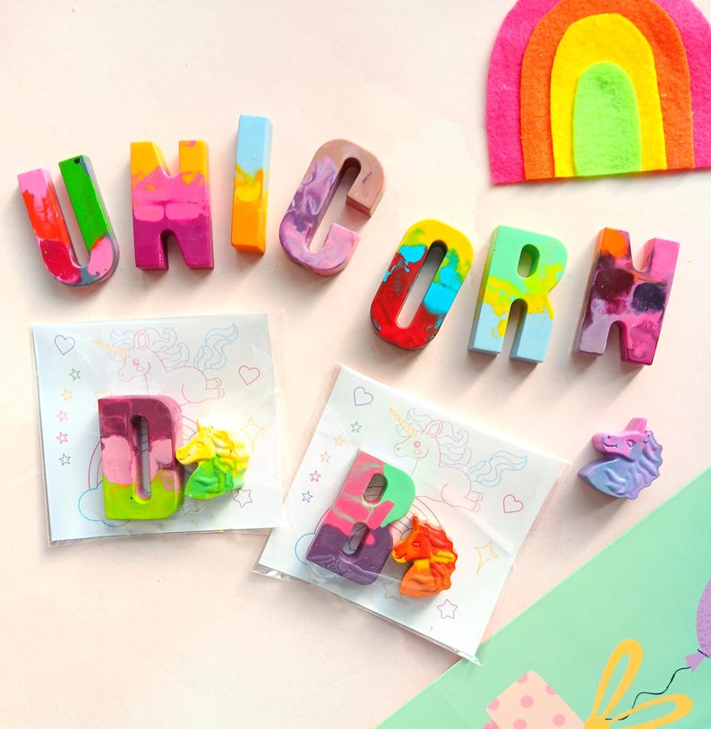 May include: A set of colorful crayon letters spelling out the word "UNICORN" with two additional crayons shaped like unicorns. The crayons are on a pink background with white paper packaging.