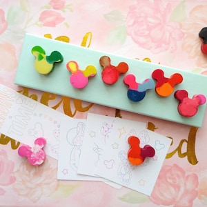 May include: A set of eight colorful Mickey Mouse shaped crayons on a light blue background. The crayons are arranged in a row and are all different colors. The text "Reading Time" is on a white bookmark with pink stars.