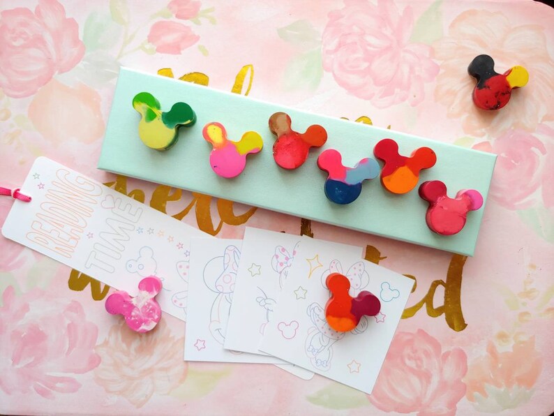 May include: A set of eight colorful Mickey Mouse shaped crayons on a light blue background. The crayons are arranged in a row and are all different colors. The text "Reading Time" is on a white bookmark with pink stars.