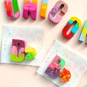 May include: A set of colorful crayon letters spelling out the word "UNICORN" with a unicorn illustration and a letter D and a unicorn crayon in a clear plastic bag.