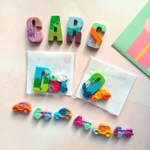 May include: A set of colorful, hand-painted crayon letters spelling out "CARS" with two smaller bags containing more crayons shaped like cars and trucks. The crayons are arranged on a pink background.