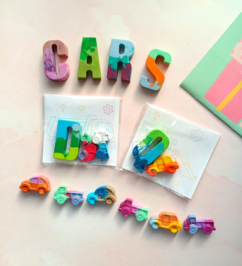 May include: A set of colorful, hand-painted crayon letters spelling out "CARS" with two smaller bags containing more crayons shaped like cars and trucks. The crayons are arranged on a pink background.