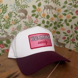 May include: White baseball cap with a burgundy brim. The cap features a rectangular patch with the words "HUMMING society" in pink lettering. The background is a floral wallpaper.