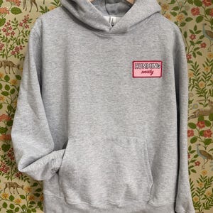 May include: A grey hoodie with a hood and a front pocket. The hoodie features a small rectangular patch on the chest with the words "HUMMING society" in white on a pink background. It is hanging on a wooden hanger.