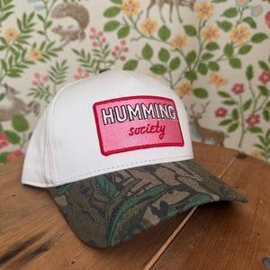 May include: White baseball cap with a camouflage brim. The cap features a pink rectangular patch with the words "HUMMING society" in white and pink lettering. The cap is sitting on a wooden surface.