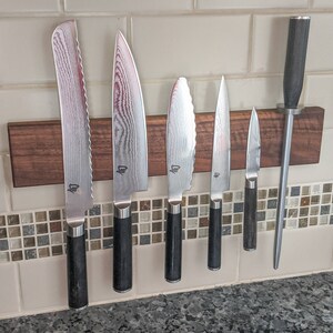 May include: A set of six kitchen knives and a honing steel attached to a wooden magnetic strip. The knives have patterned blades and black handles. The honing steel has a black handle and a silver steel rod.