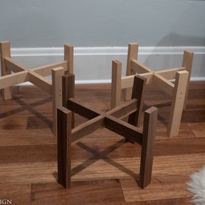 May include: Four wooden plant stands in varying shades of brown and natural wood tones. The stands have a cross-shaped design with four vertical legs. The stands are displayed on a hardwood floor.