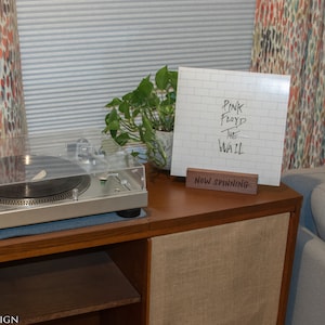 May include: A silver turntable with a clear cover sits on a wooden cabinet. A vinyl record with the album cover "Pink Floyd The Wall" is displayed on a wooden stand that reads "Now Spinning". A green plant is next to the record.