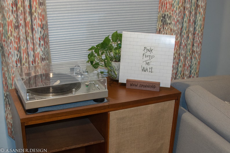 May include: A silver turntable with a clear cover sits on a wooden cabinet. A vinyl record with the album cover "Pink Floyd The Wall" is displayed on a wooden stand that reads "Now Spinning". A green plant is next to the record.