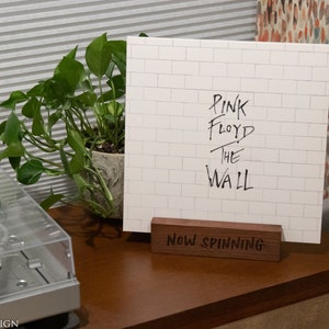 May include: A white album cover with the text "PINK FLOYD THE WALL" displayed on a wooden stand that reads "Now Spinning." The album cover is propped up on a wooden stand on a wooden surface, next to a turntable and a potted plant.