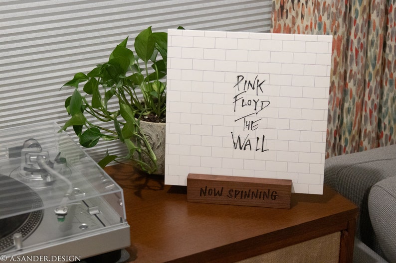 May include: A white album cover with the text "PINK FLOYD THE WALL" displayed on a wooden stand that reads "Now Spinning." The album cover is propped up on a wooden stand on a wooden surface, next to a turntable and a potted plant.