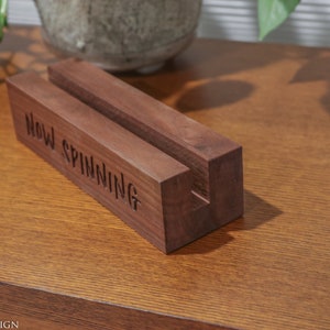 May include: A wooden record stand with the words "Now Spinning" carved into the front. The stand is a rich brown color and has a slot to hold a record. The stand is on a wooden surface.