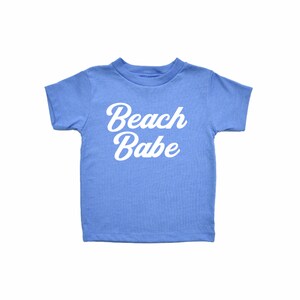 May include: A light blue t-shirt with the words "Beach Babe" in white script. The shirt has short sleeves and a crew neck, perfect for a day at the beach or a casual summer outfit.