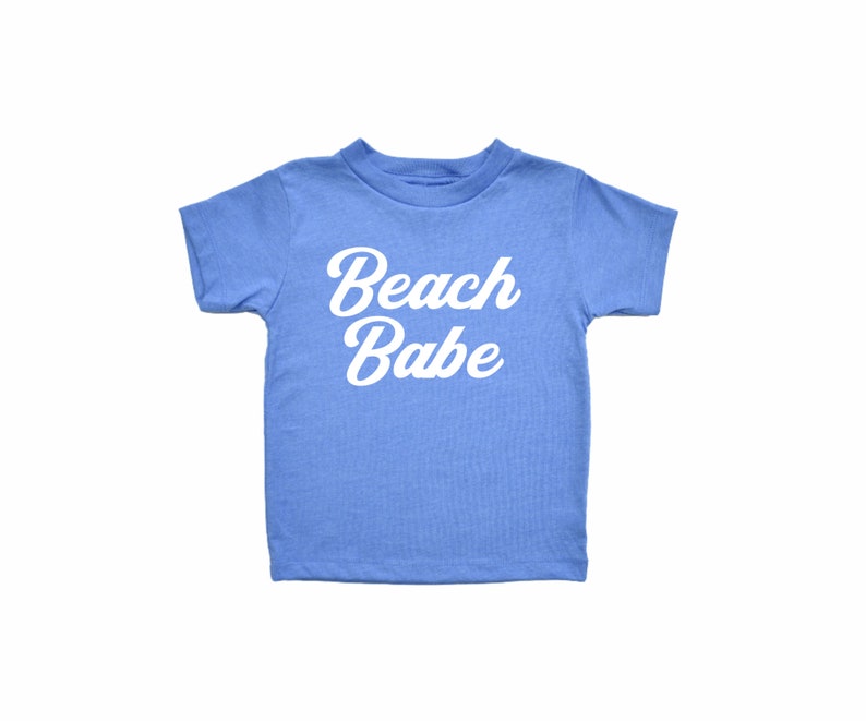 May include: A light blue t-shirt with the words "Beach Babe" in white script. The shirt has short sleeves and a crew neck, perfect for a day at the beach or a casual summer outfit.