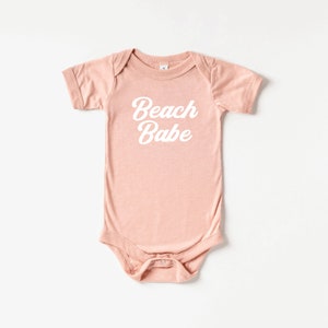 May include: A short-sleeved, light pink baby bodysuit with the words "Beach Babe" printed in white script across the front. The bodysuit has a snap closure at the bottom and a simple, classic design.