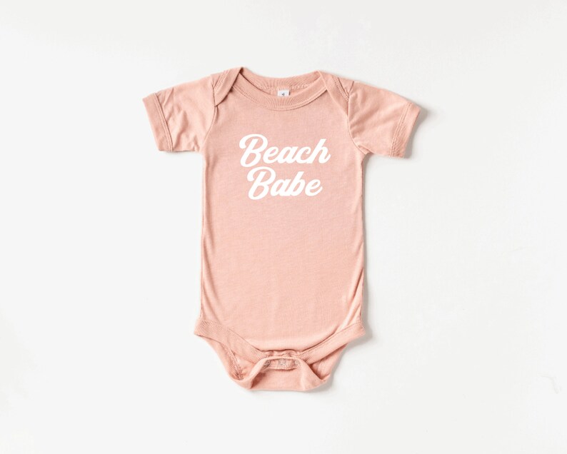 May include: A short-sleeved, light pink baby bodysuit with the words "Beach Babe" printed in white script across the front. The bodysuit has a snap closure at the bottom and a simple, classic design.