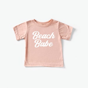 May include: A light pink t-shirt with the words "Beach Babe" in white script. The short-sleeved shirt is made of a soft material and is suitable for children. The shirt is a casual wear item.