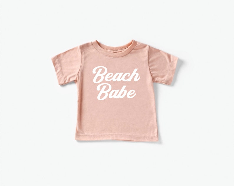 May include: A light pink t-shirt with the words "Beach Babe" in white script. The short-sleeved shirt is made of a soft material and is suitable for children. The shirt is a casual wear item.