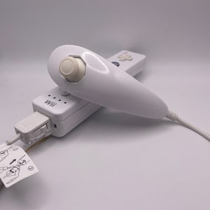 May include: A white Nintendo Wii remote with a white Wii MotionPlus attachment. The remote is plugged into a white Wii console. The text "Wii" is visible on the console.