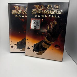 May include: Two copies of the "Dead Space Downfall" DVD. The cover art features a black gloved hand reaching out against a fiery background. The title is in gold lettering. A sticker on the front reads "Prequel to the Dead Space Game From EA."