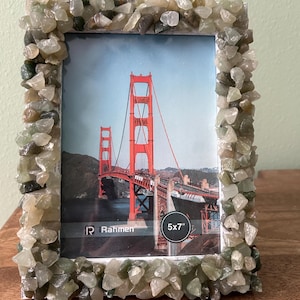 May include: A rectangular picture frame decorated with small, polished green and translucent stones. The frame holds a photograph of the Golden Gate Bridge in San Francisco. The text "Rahmen" and "5x7"" are printed on the lower portion of the image.