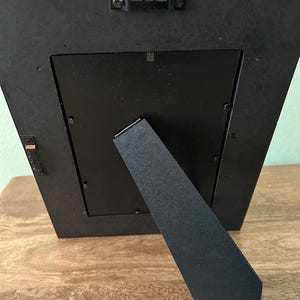 May include: A black rectangular picture frame shown from the back, featuring a central easel stand, metal swivel clips to secure the backing, and two metal sawtooth hangers attached to the frame edge for both vertical and horizontal wall mounting.