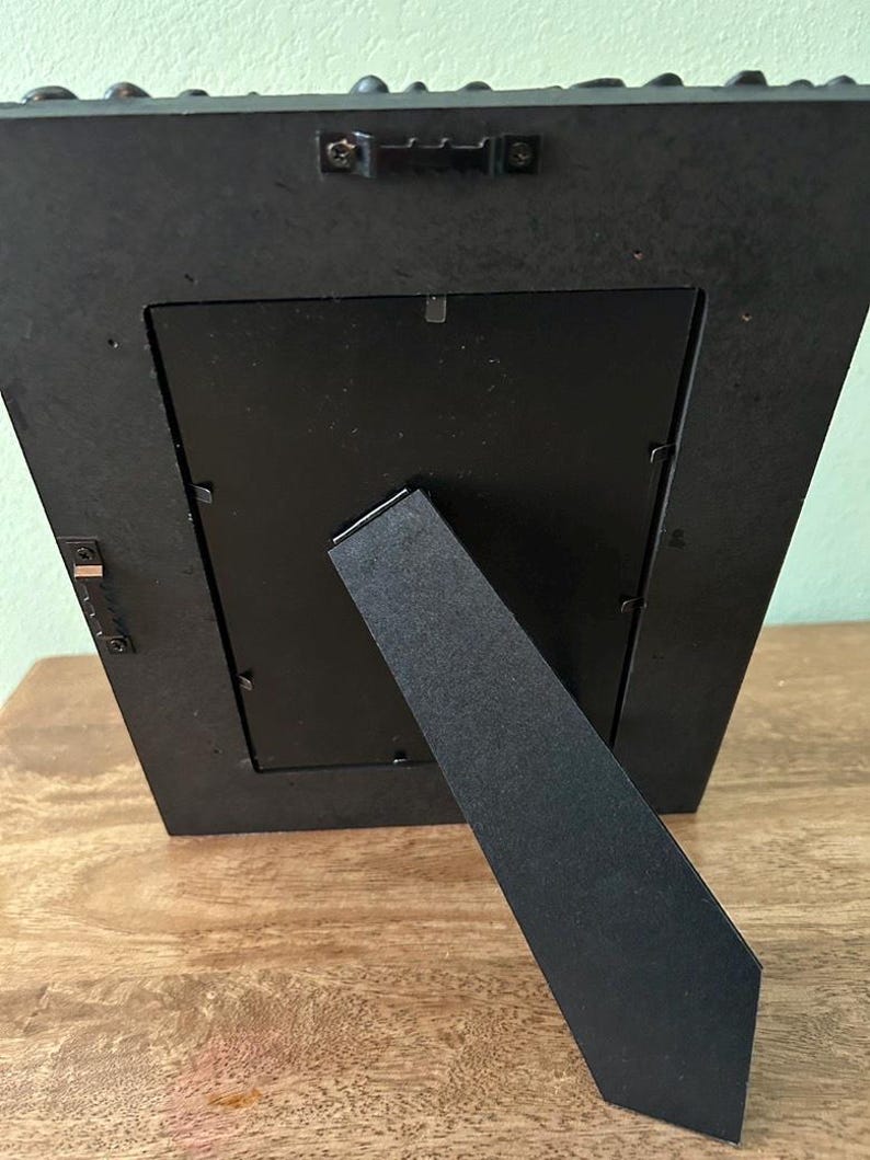 May include: A black rectangular picture frame shown from the back, featuring a central easel stand, metal swivel clips to secure the backing, and two metal sawtooth hangers attached to the frame edge for both vertical and horizontal wall mounting.