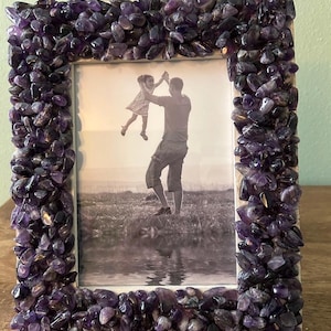 May include: A rectangular picture frame featuring a border densely covered in small, polished purple amethyst stones. The frame holds a black and white photograph of an adult lifting a child into the air outdoors near a body of water.