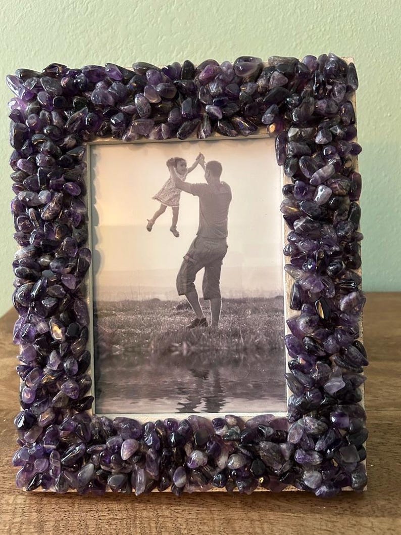 May include: A rectangular picture frame featuring a border densely covered in small, polished purple amethyst stones. The frame holds a black and white photograph of an adult lifting a child into the air outdoors near a body of water.